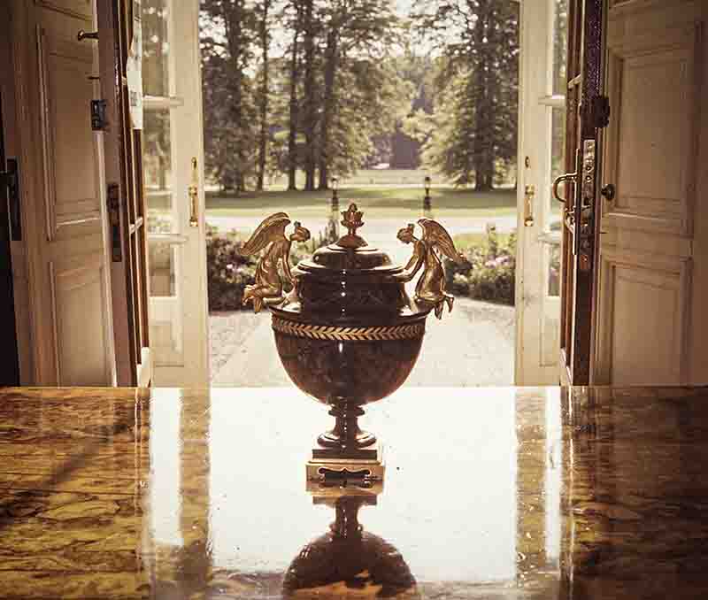 Eine Fotografie von Peter Bock-Schroeder aus dem Jahr 1960, aufgenommen im Haus Doorn. Im Zentrum steht eine prunkvolle Prunkvase aus dunklem Stein mit vergoldeten Bronzemontierungen und Engelsfiguren auf einem hochglanzpolierten Tisch. Durch eine weit geöffnete Fenstertür im Hintergrund fällt der Blick auf den herrschaftlichen Park des Schlosses.